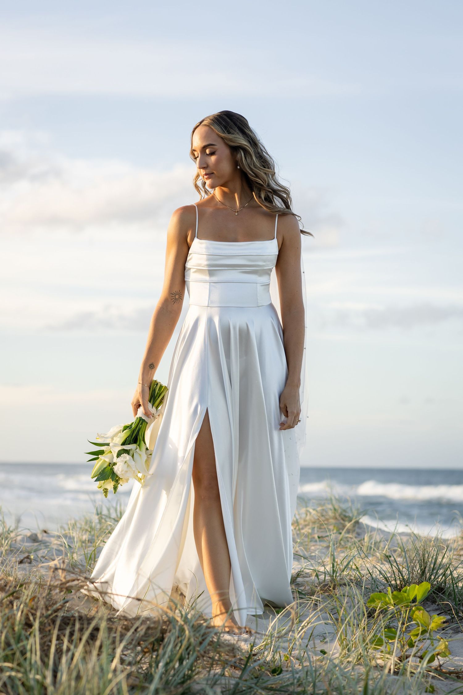 Woman in flowing white dress stands on coastal dunes holding flowers at sunset.beach wedding currumbin surf club wedding