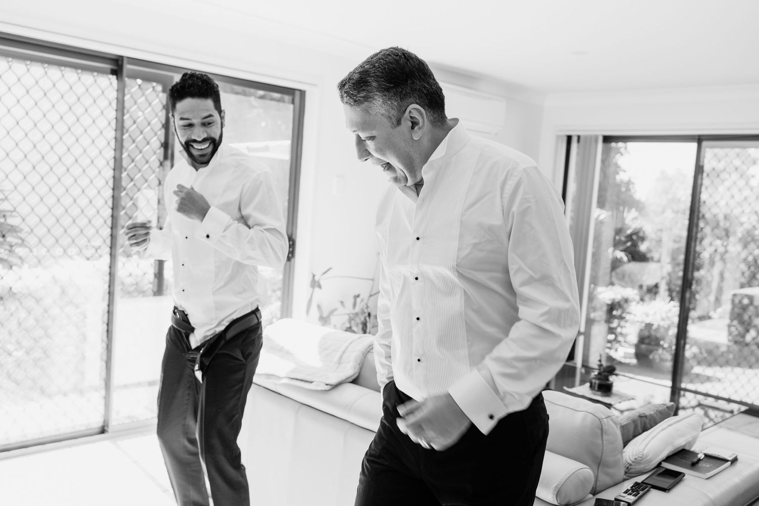 Two men in formal attire share a lighthearted moment while getting ready in a bright indoor space.