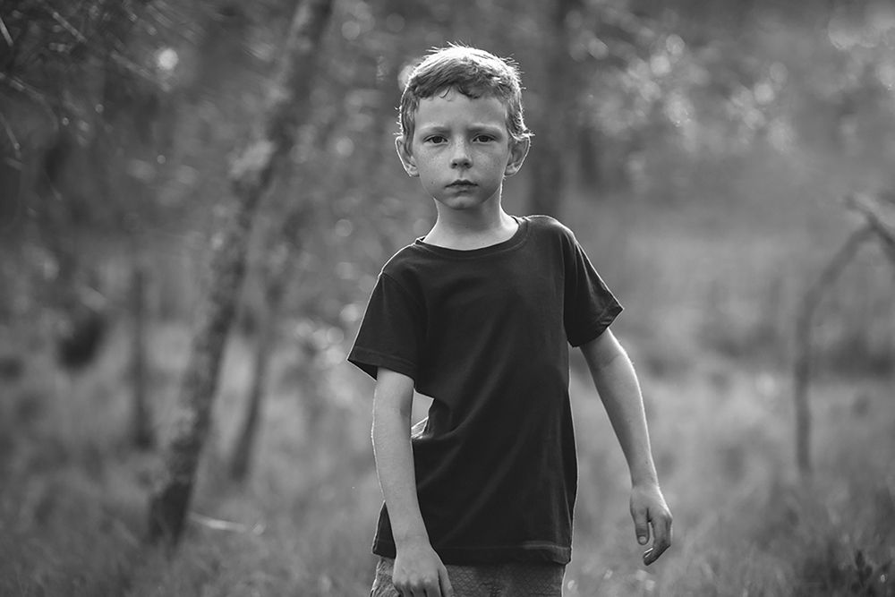 Black and white portrait of a young boy in a dark t-shirt standing outdoors in a wooded area.