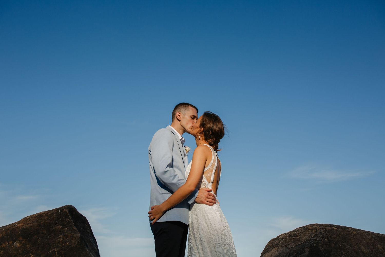 Newlyweds share intimate moment on rocky cliff against vibrant blue sky.Gold Coast beach wedding photography. Froggies beach
