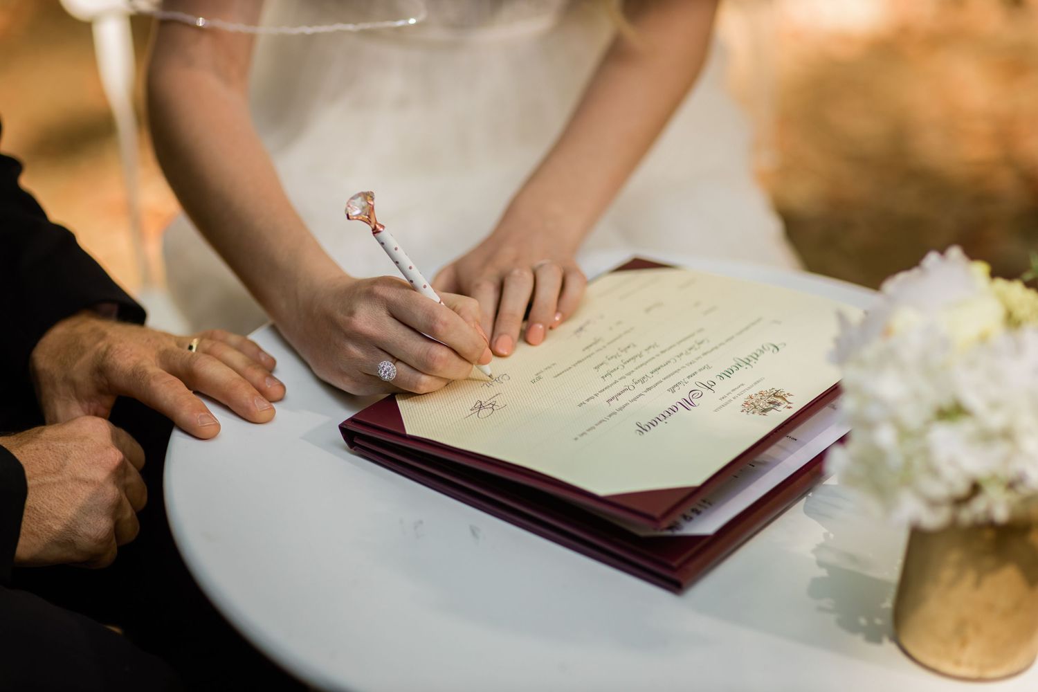 Close-up of hands signing marriage certificate with white flowers on table.Currumbin Valley wedding photography Gold Coast