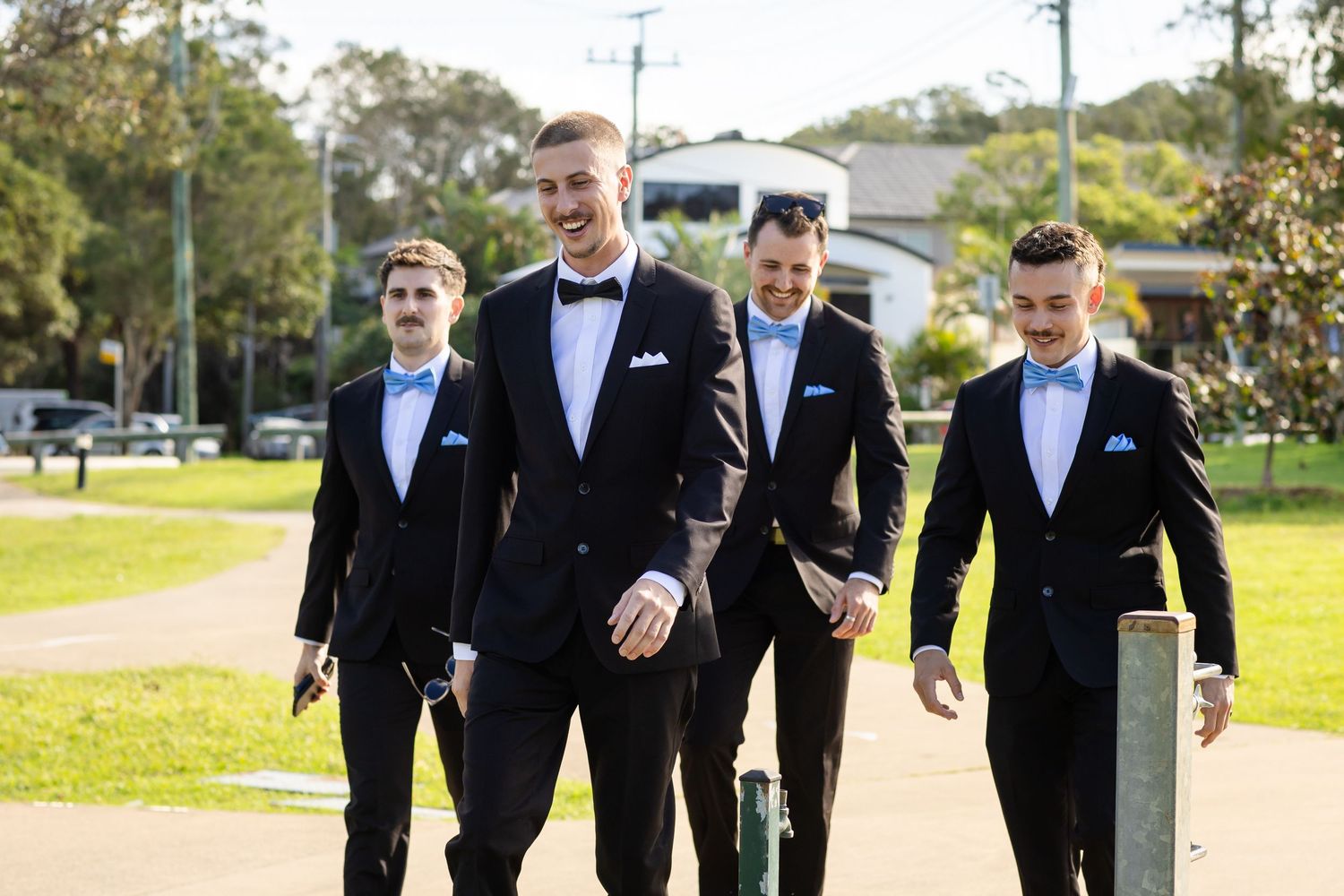 Groomsmen in black tuxedos walk together outdoors on a sunny day.beach wedding currumbin surf club wedding