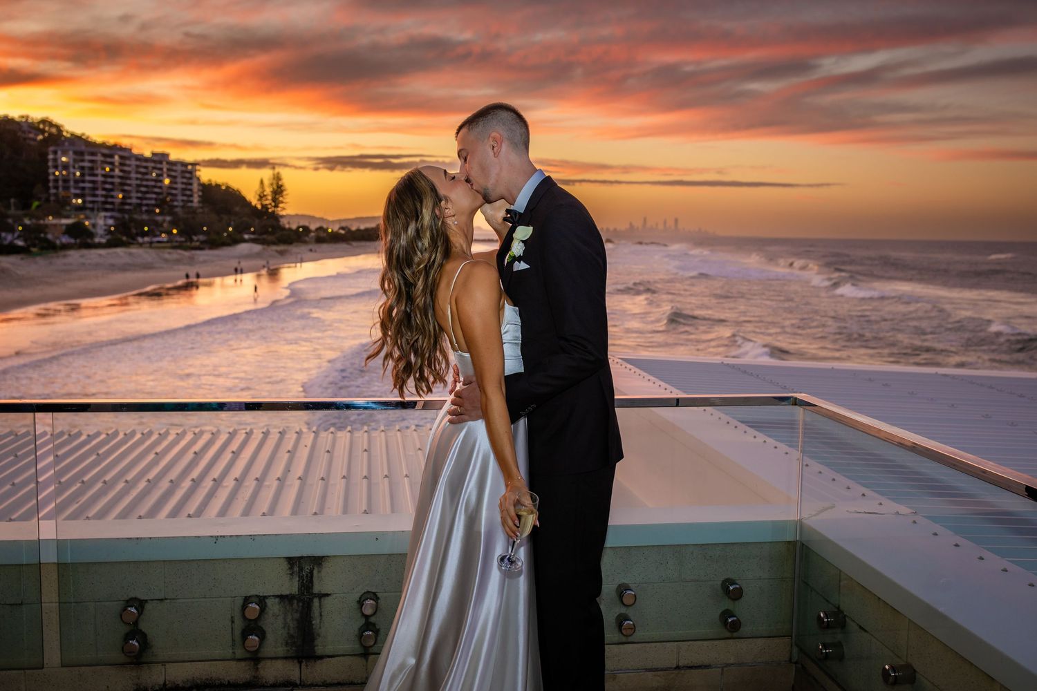 Couple shares a romantic moment on a balcony during sunset with dramatic orange sky.beach wedding currumbin surf club wedding