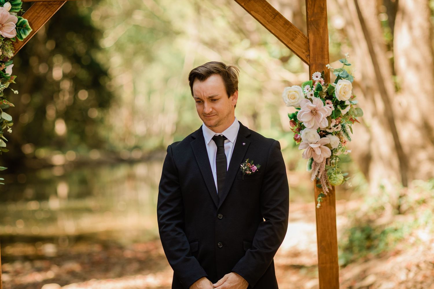 Outdoor wedding ceremony in a sunlit wooded area with a rustic wooden arch decorated with floral arrangements.Currumbin Valley wedding photo