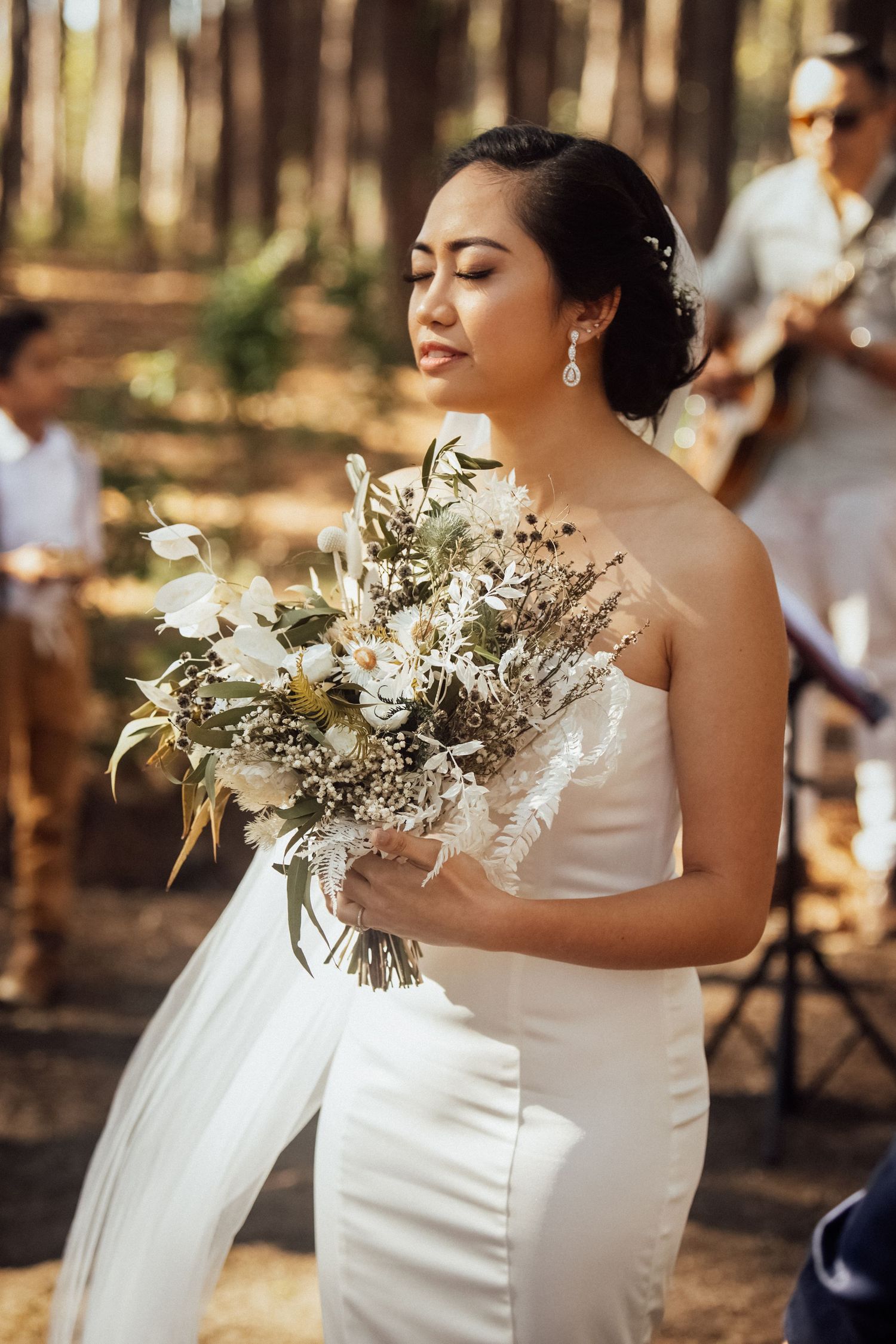Bride holding a delicate bouquet of white and dried flowers against a forest backdrop.pizzey park wedding Gold Coast