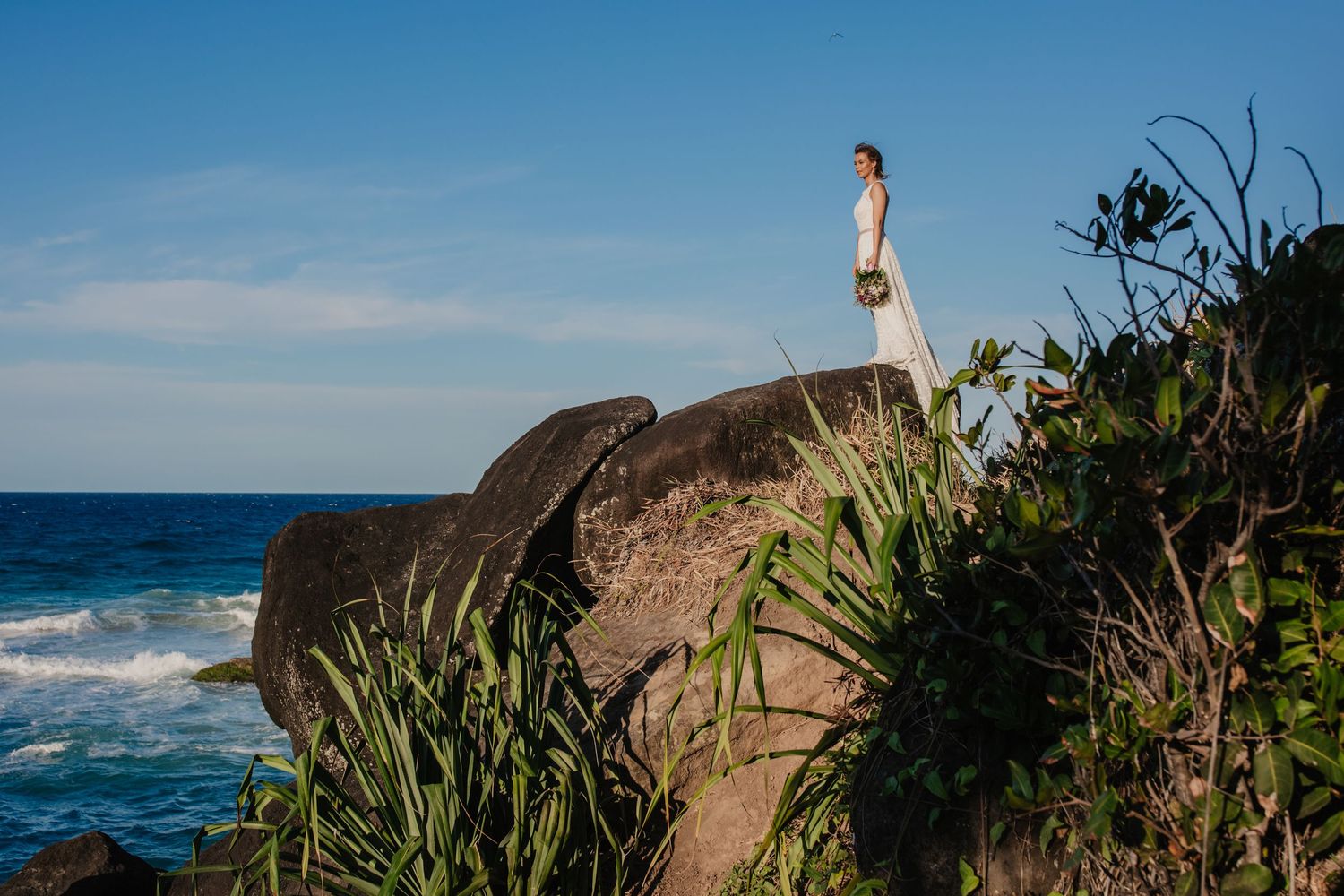 Bride in white dress stands alone on dramatic oceanside cliff with waves crashing below.Gold Coast beach wedding photography. Froggies beach