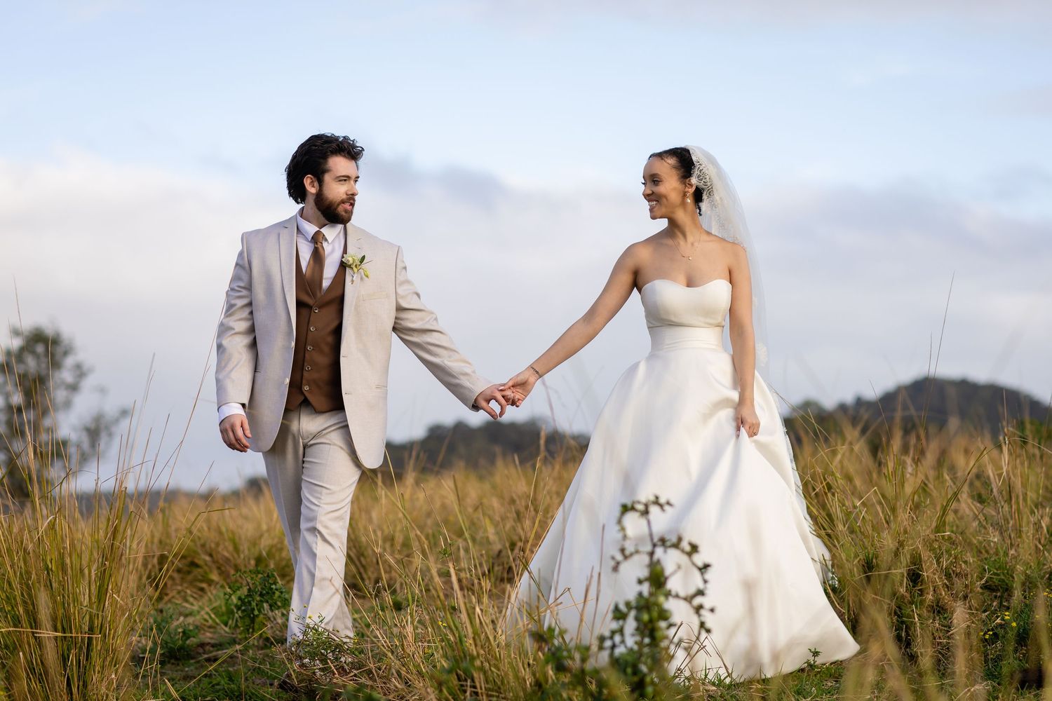 Couple walking hand in hand through a scenic field at sunset in formal wedding attire.The barrow restaurant mount Nathan wedding