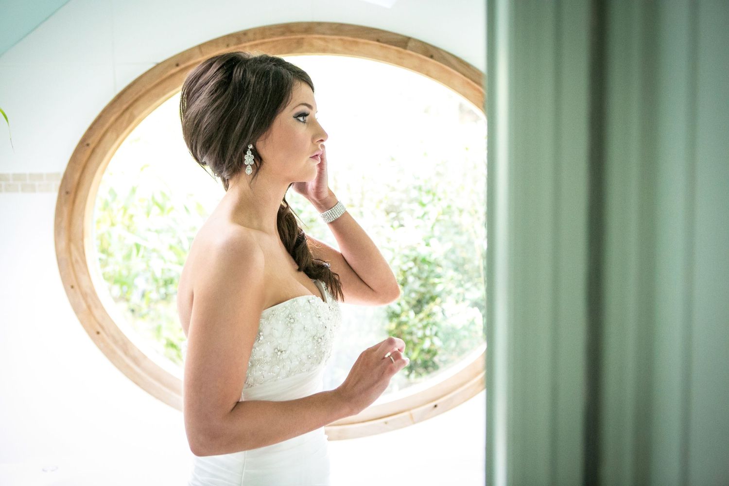 Bride in white dress framed by circular window with soft natural lighting. Bridal prep photos inspo.
