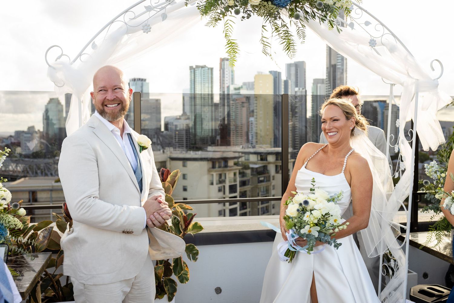 Elegant white wedding arch decorated with greenery and flowers against a city skyline backdrop.wedding ceremony at iris rooftop hotel in bri