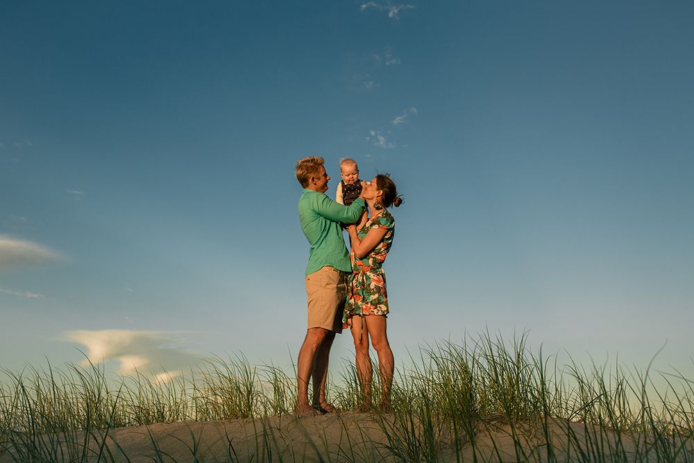 A family of three stands on sandy dunes with green grass, sharing a joyful moment under a blue sky at sunset.