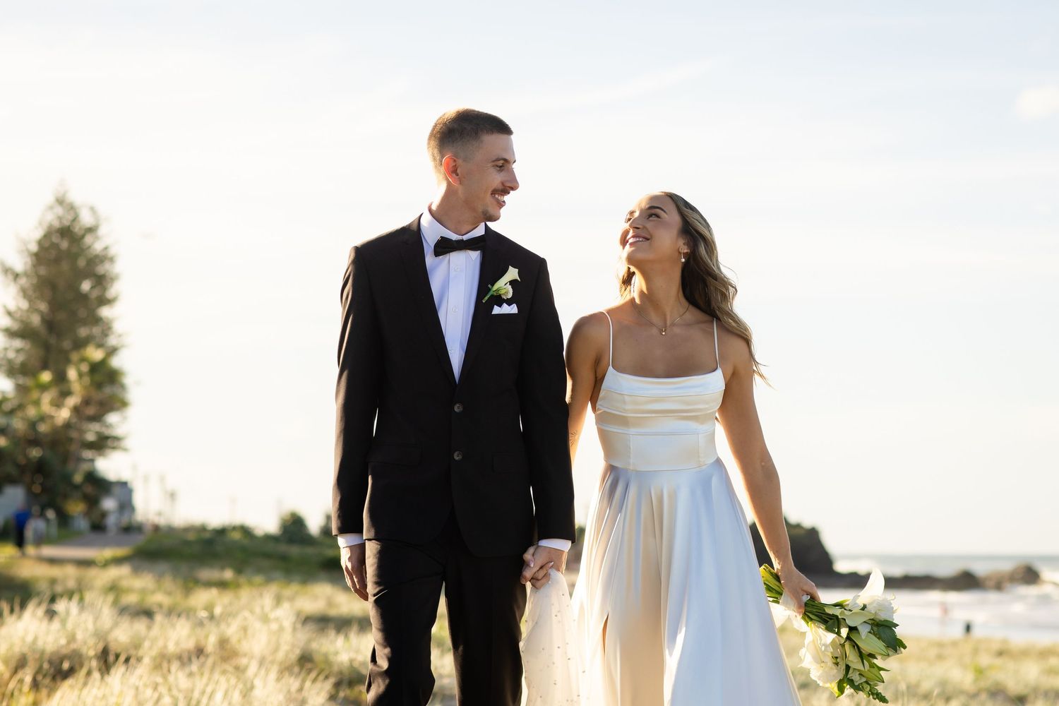 Newlyweds walk hand in hand through a sunlit coastal landscape.beach wedding currumbin surf club wedding