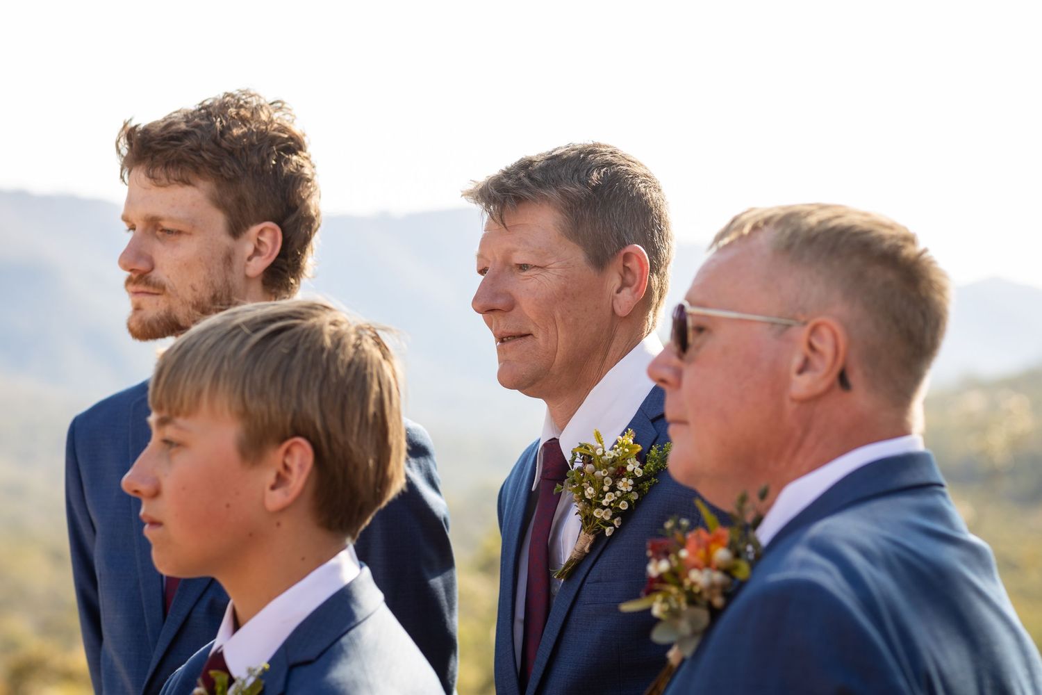 Groomsmen in navy suits and plaid accessories stand in line during outdoor ceremony.