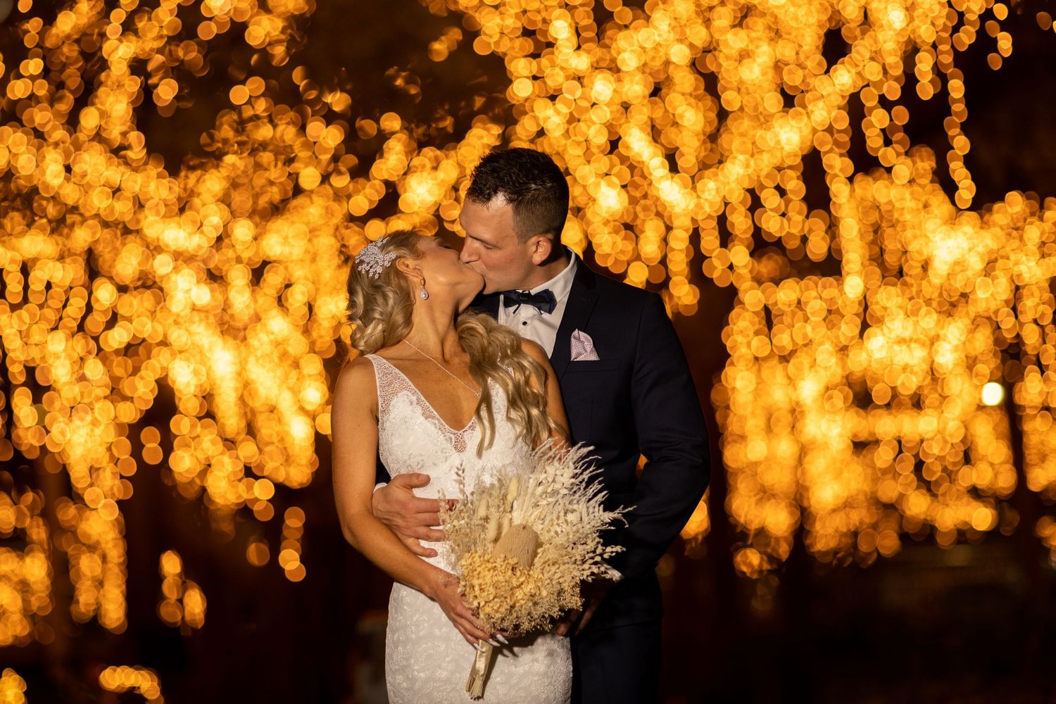 A couple shares a romantic kiss under glowing golden string lights at a nighttime wedding celebration.
