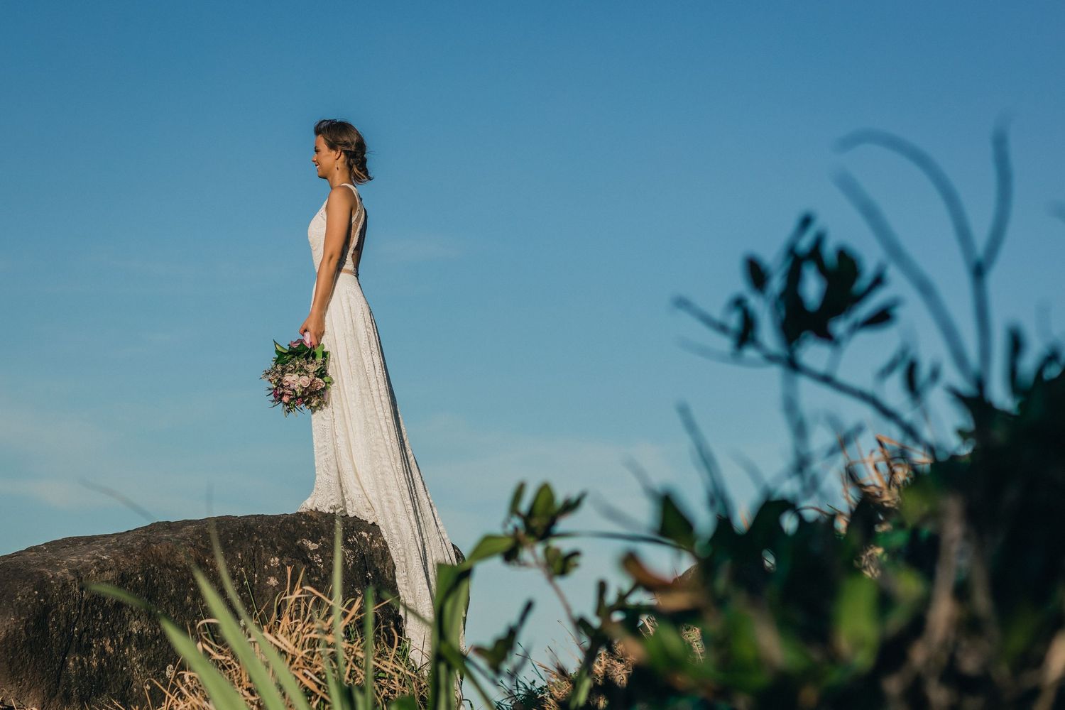 A bride in a flowing white dress stands on a hilltop against a bright blue sky holding a bouquet.Gold Coast beach wedding photography. Frogg