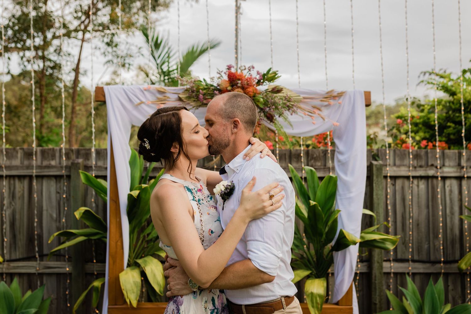 Couple embracing under a white arch decorated with tropical flowers and greenery.Gold Coast elopement photographer