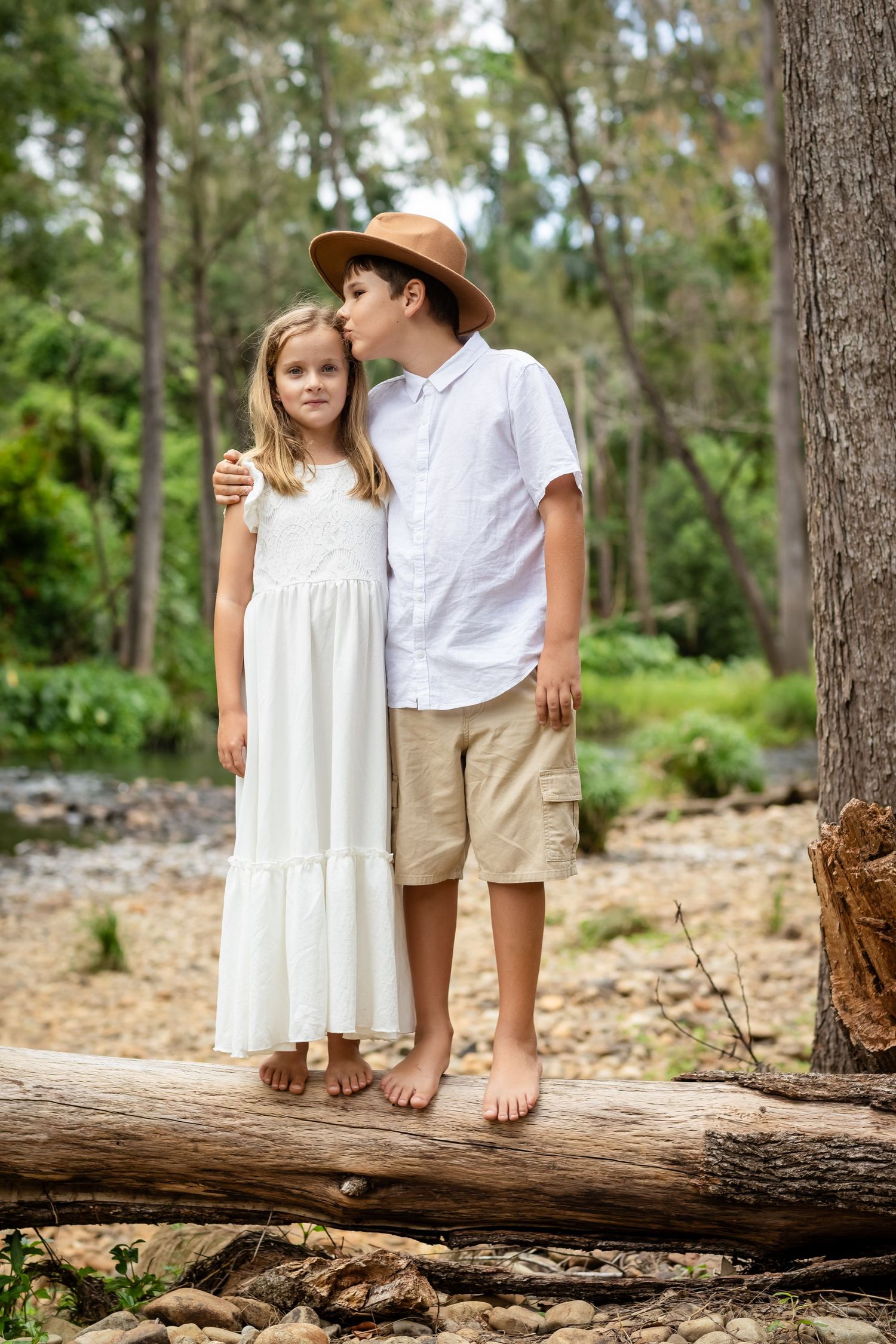Two children in light summer clothing stand barefoot on a fallen tree trunk in a sunlit forest.