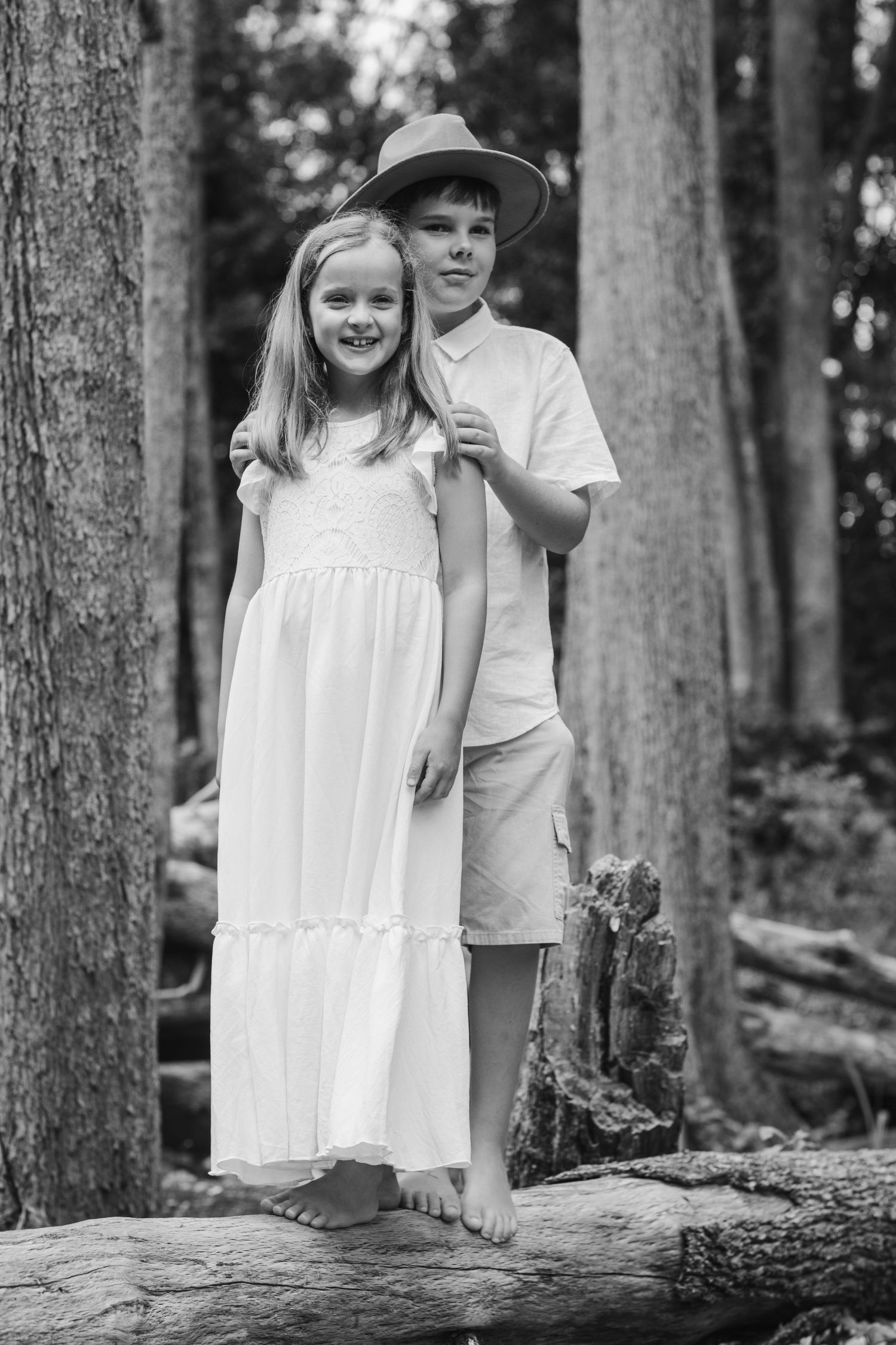 A black and white portrait of two siblings standing together on a fallen log in a wooded forest setting.Tallebudgera family photography. Gol