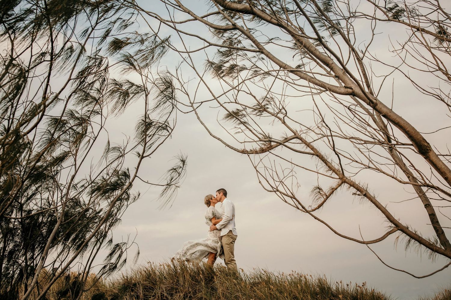 A romantic couple embraces on a hilltop surrounded by bare tree branches against a cloudy sky at sunset.