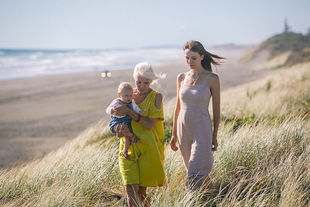 Three generations of women, including a baby, stand among beach dunes with ocean waves in the background.