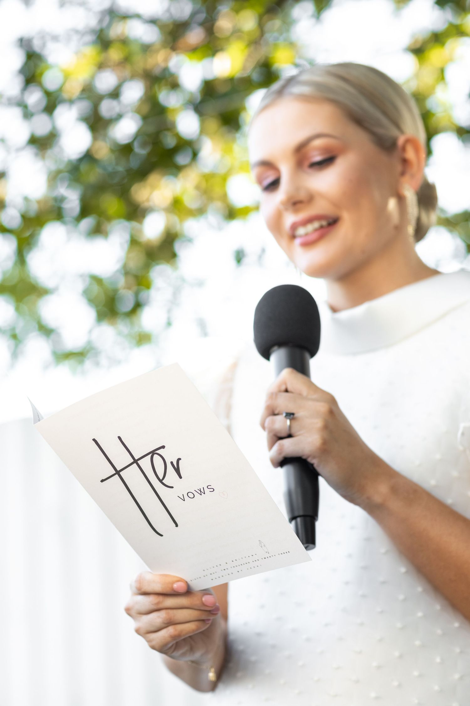 Wedding vows being read outdoors with a microphone against a leafy background.