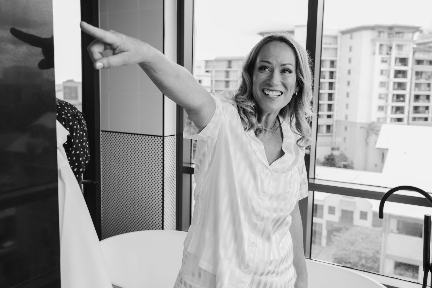 Woman in white robe pointing excitedly on balcony with urban backdrop.