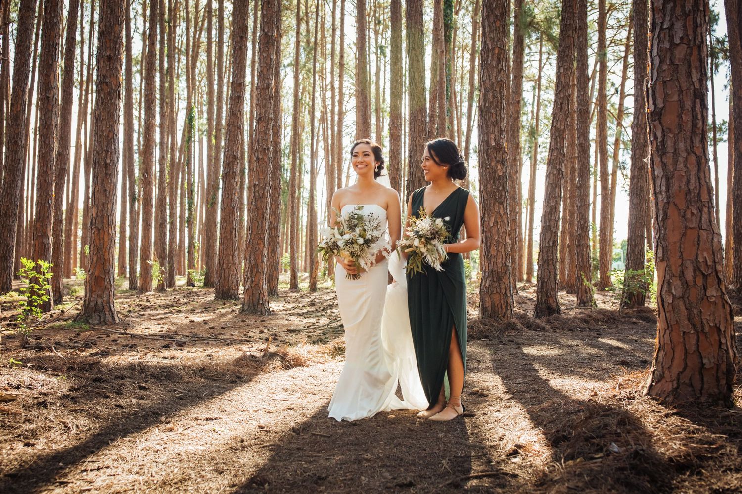 Two people in formal attire stand together holding bouquets in a sunlit pine forest.