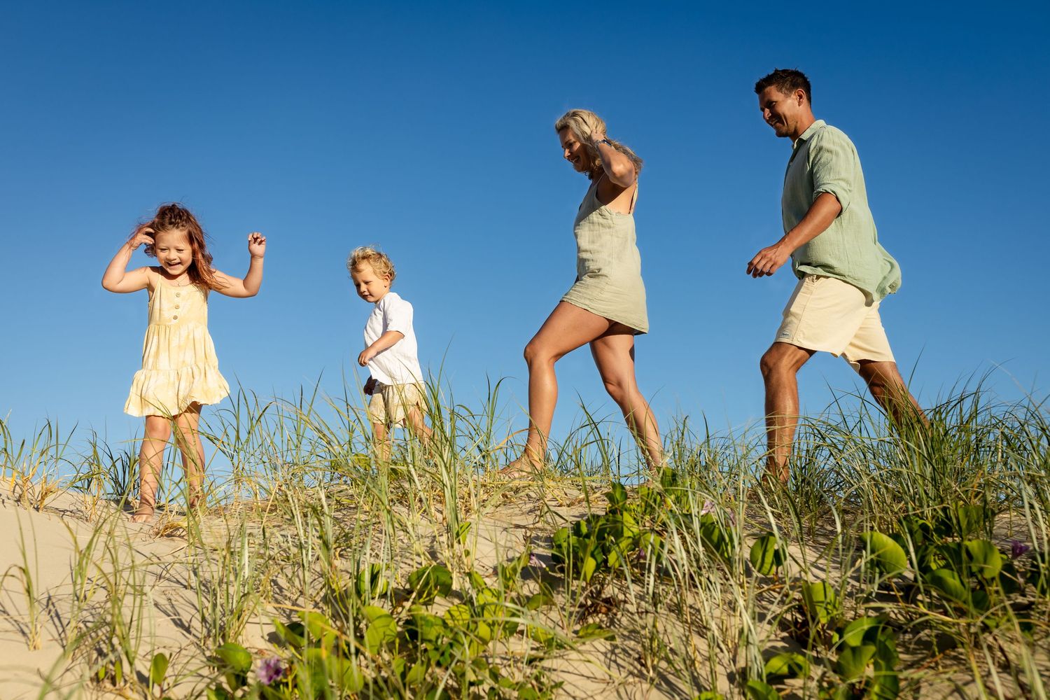 People walking along grassy sand dunes against bright blue sky.Family photography Kirra beach Gold Coast.Gold Coast candid family photograph
