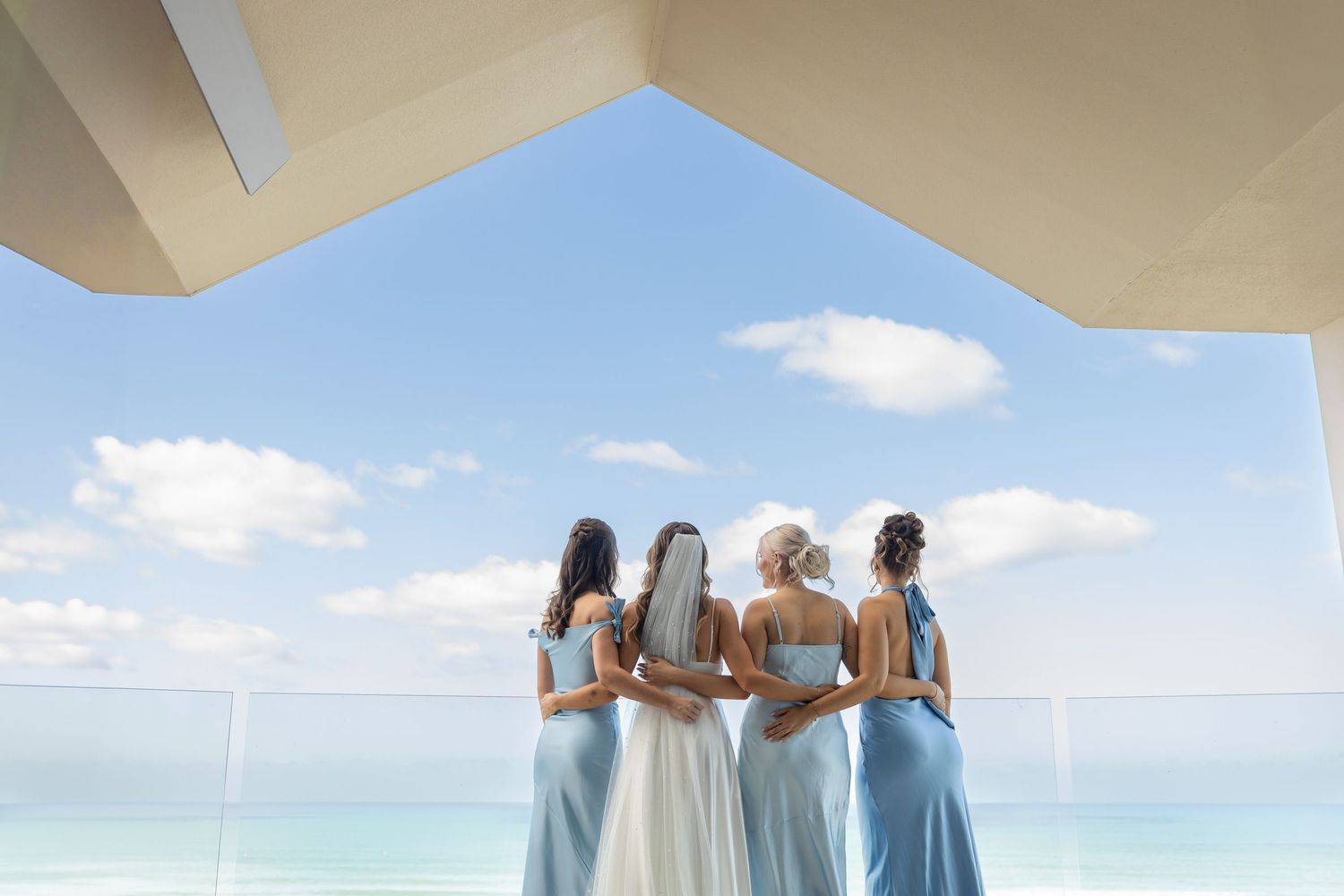 Bridesmaids in light blue dresses stand with bride overlooking ocean view on sunny day.candid wedding photography Gold Coast