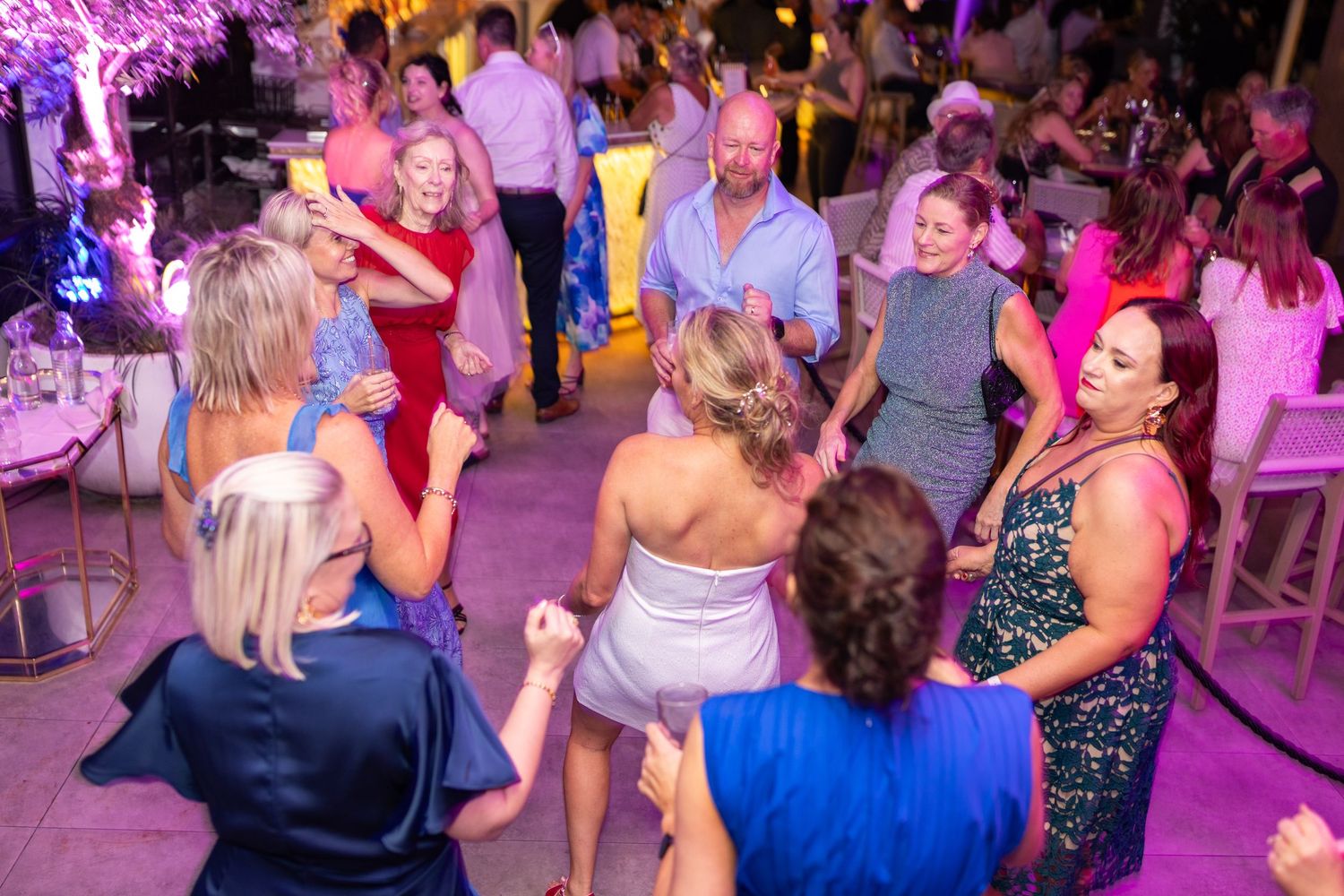 Wedding party dancing under purple mood lighting at an indoor reception venue.