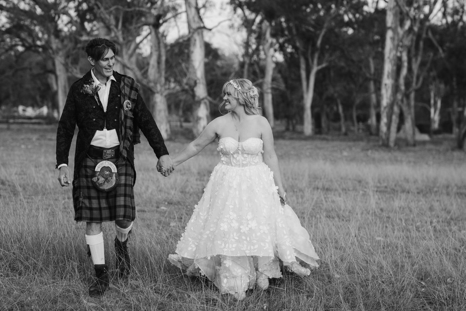 A couple in formal attire walking together through a grassy outdoor area.