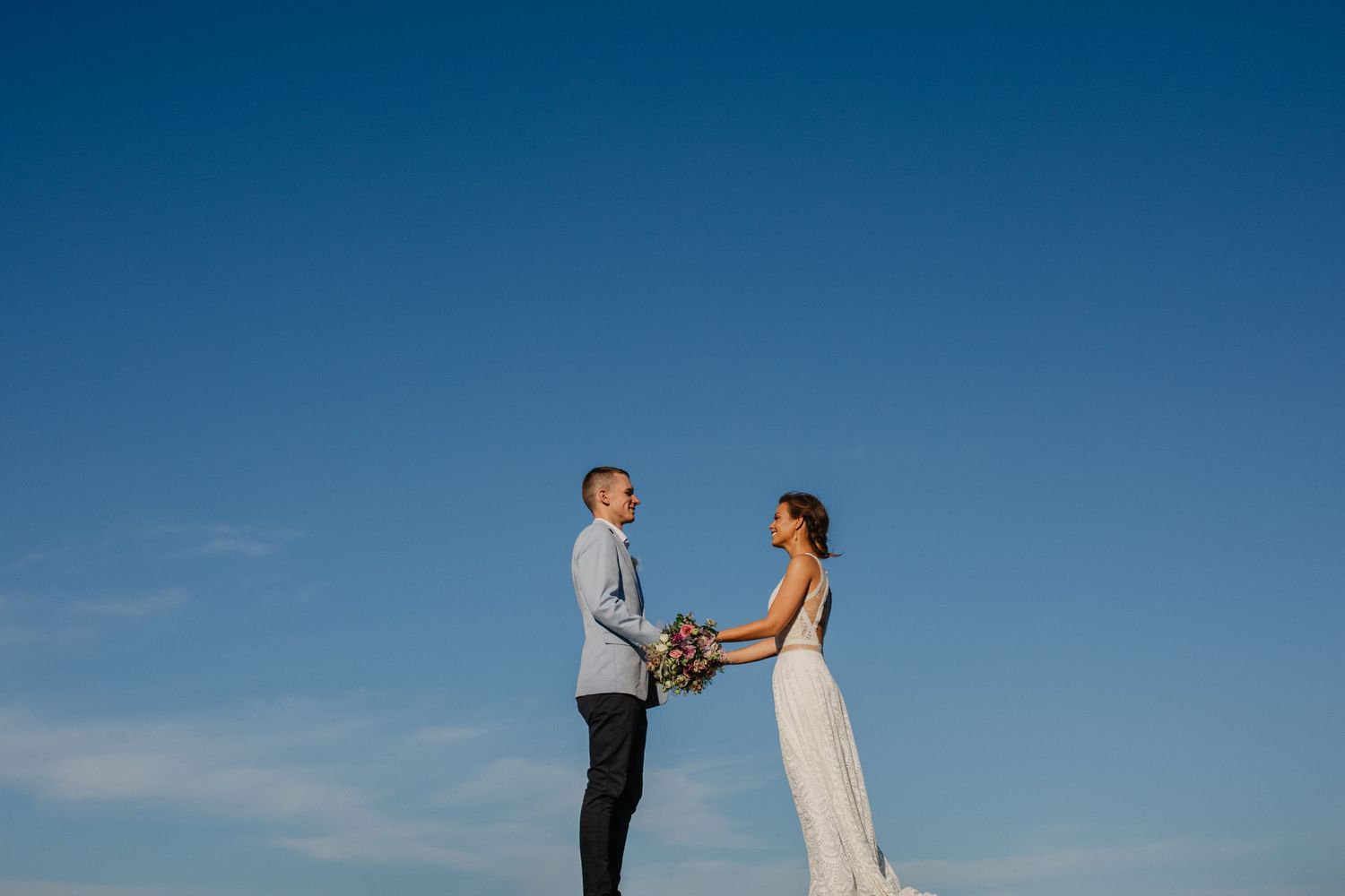 Couple holds hands while standing against bright blue sky during wedding ceremony.Gold Coast beach wedding photography. Froggies beach