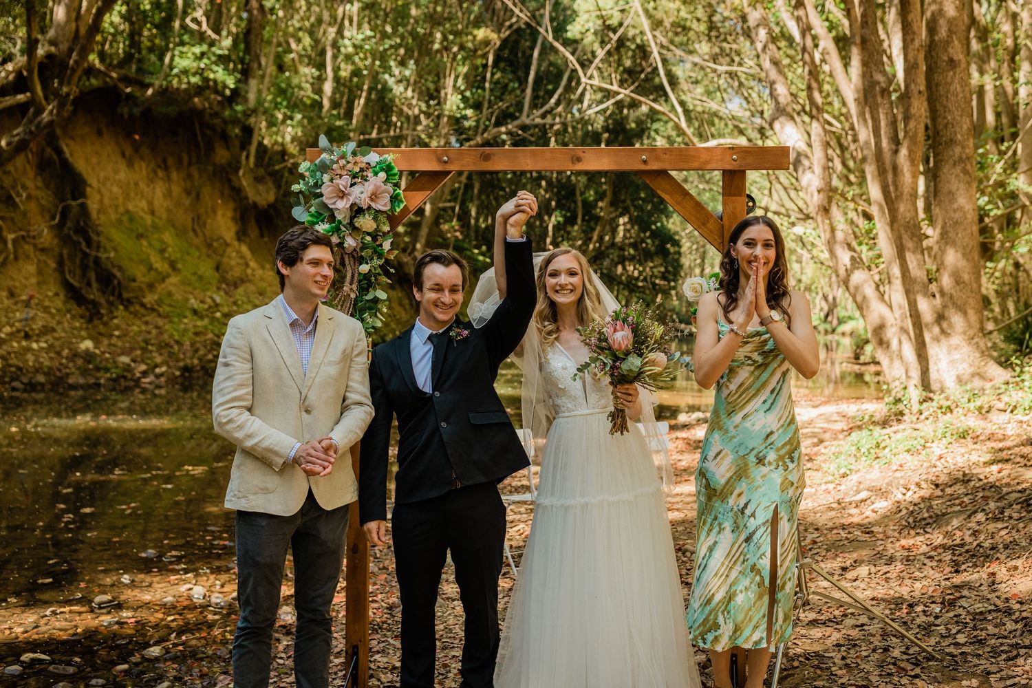Wedding party celebrating under wooden arch in forest setting.Currumbin Valley wedding photography Gold Coast