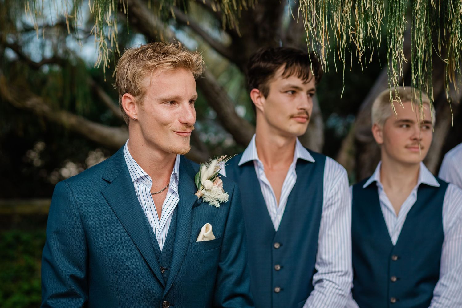 Groomsmen in navy suits and white shirts stand together during an outdoor wedding ceremony.