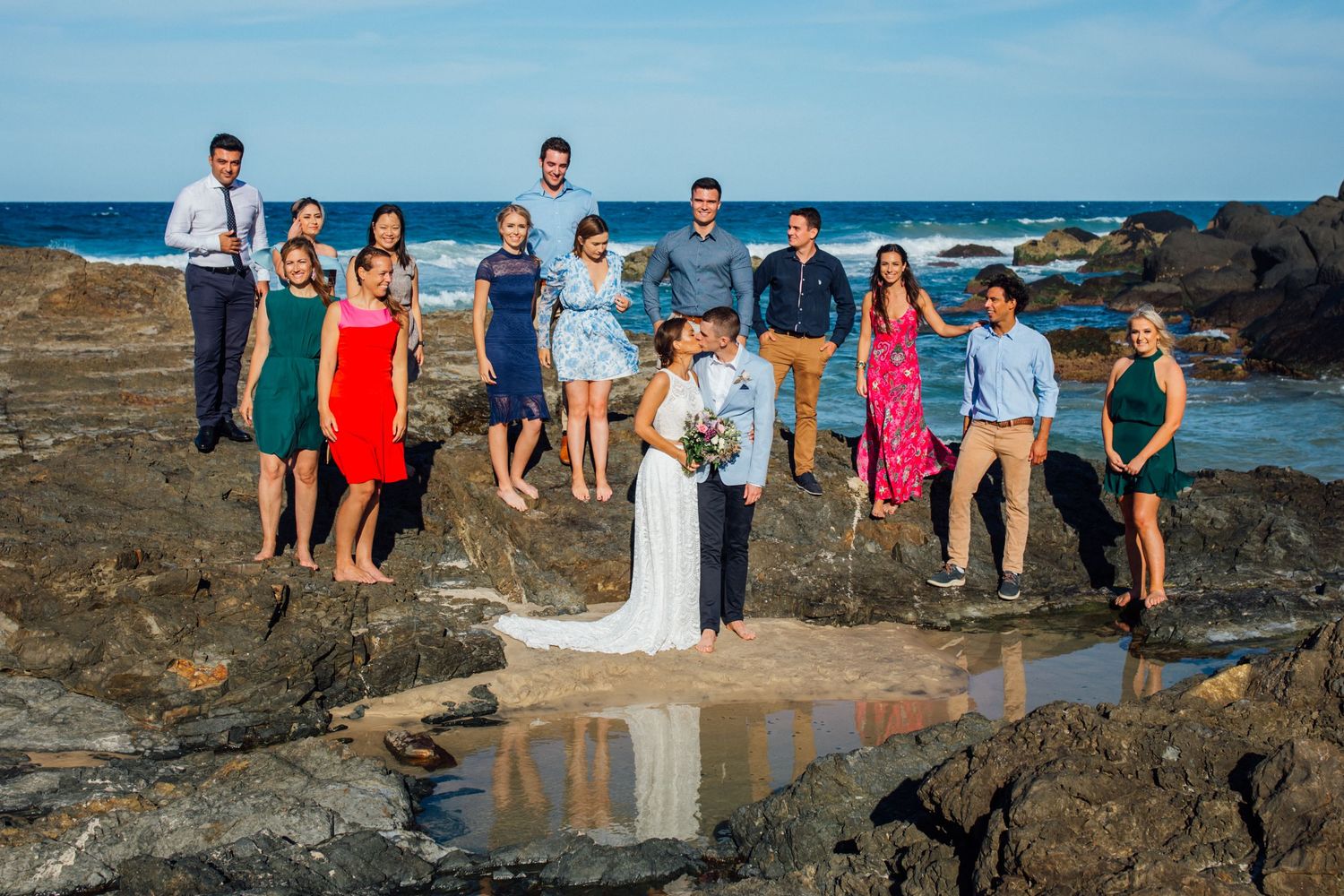 Wedding party poses on rocky coastal outcrop with ocean and blue sky in background during sunset celebration.Gold Coast beach wedding photog