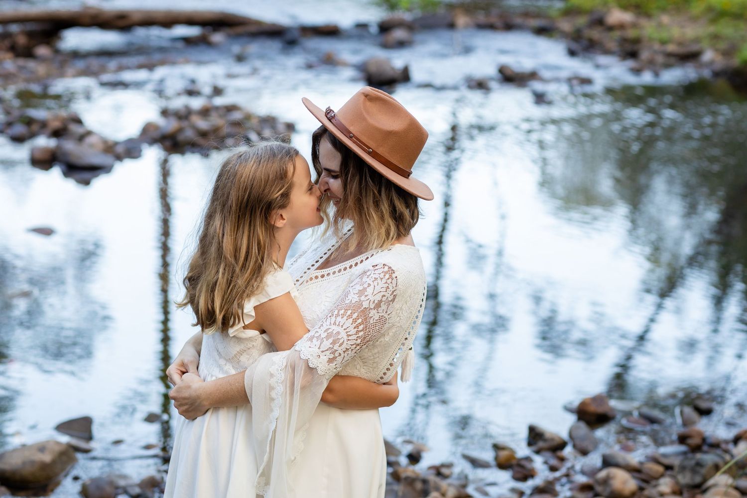 Two people in white dresses embrace by a reflective stream with rocks. Tallebudgera family photography. Gold Coast family photographer