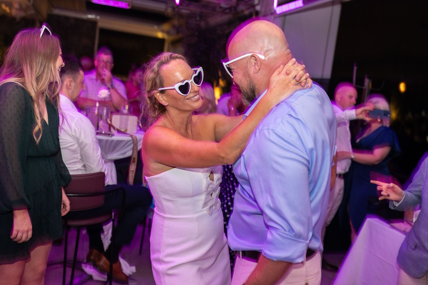 Wedding guests dancing together under vibrant purple lighting at evening reception.