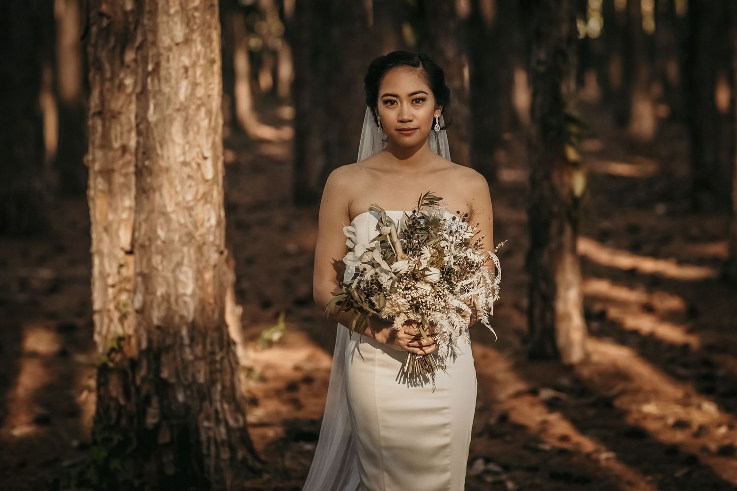 Elegant bride holds dried flower bouquet while standing among tall pine trees.pizzey park wedding Gold Coast