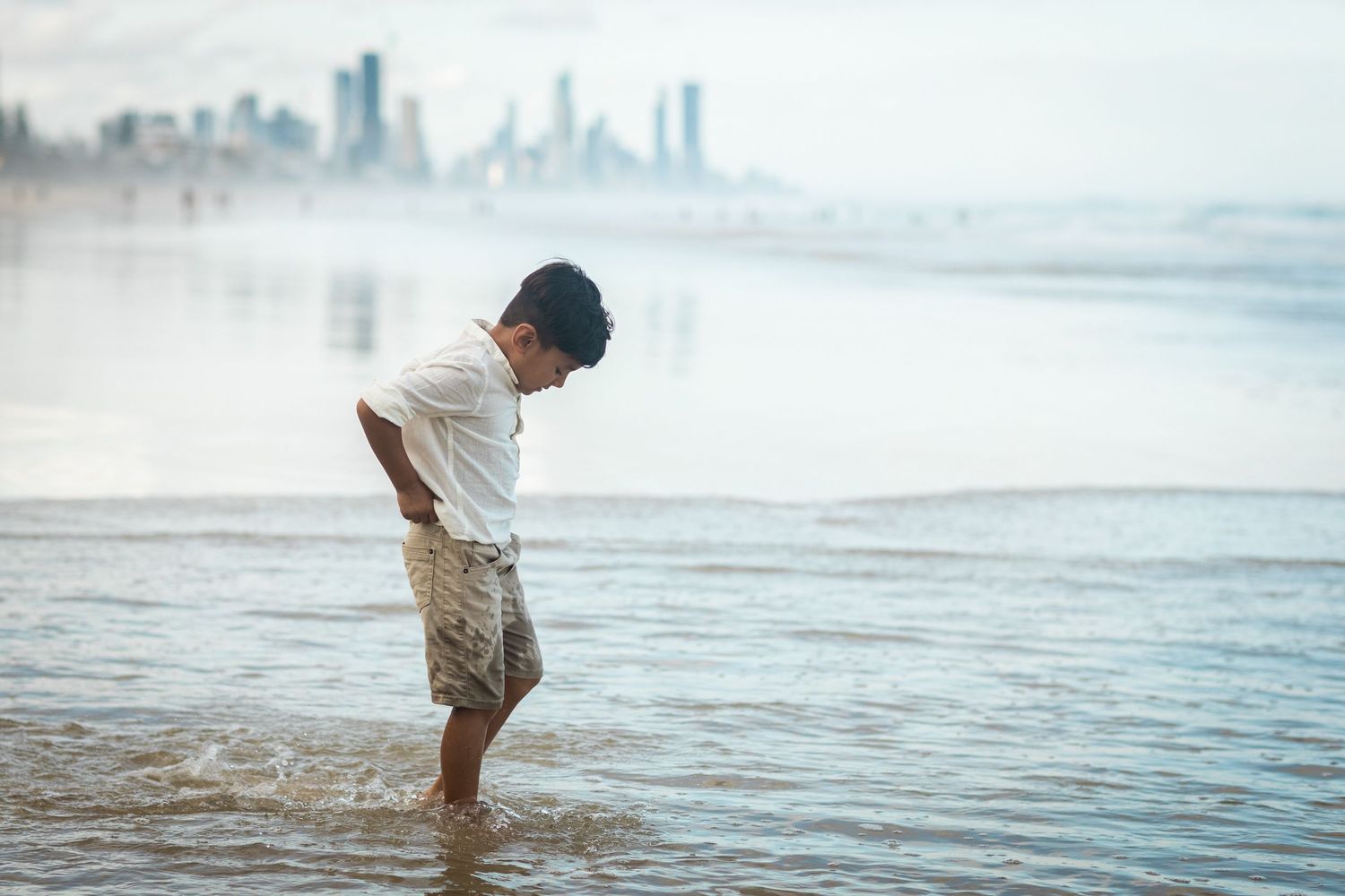Child playing in shallow ocean water with city skyline visible in background.Gold Coast candid family photographer.