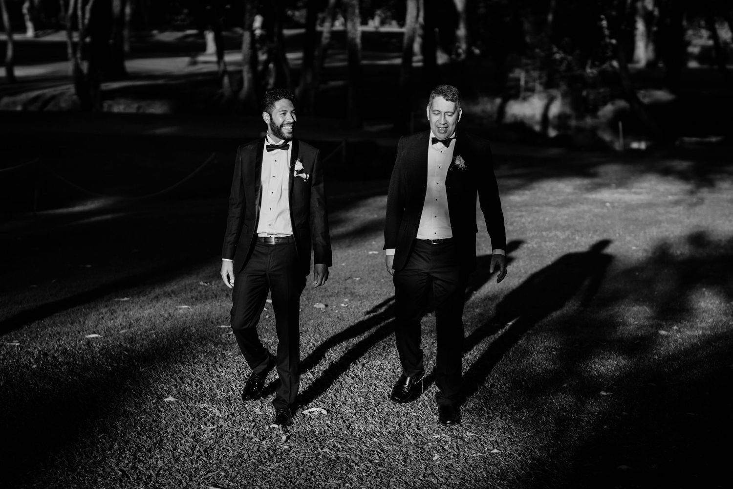 Two men in formal tuxedos walking together on a gravel path in dramatic black and white lighting. Surfers Paradise Golf Club wedding.