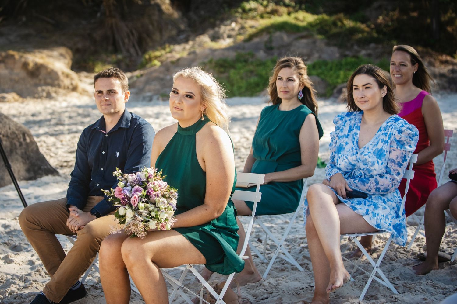 Wedding guests seated casually on beach rocks during outdoor ceremony.