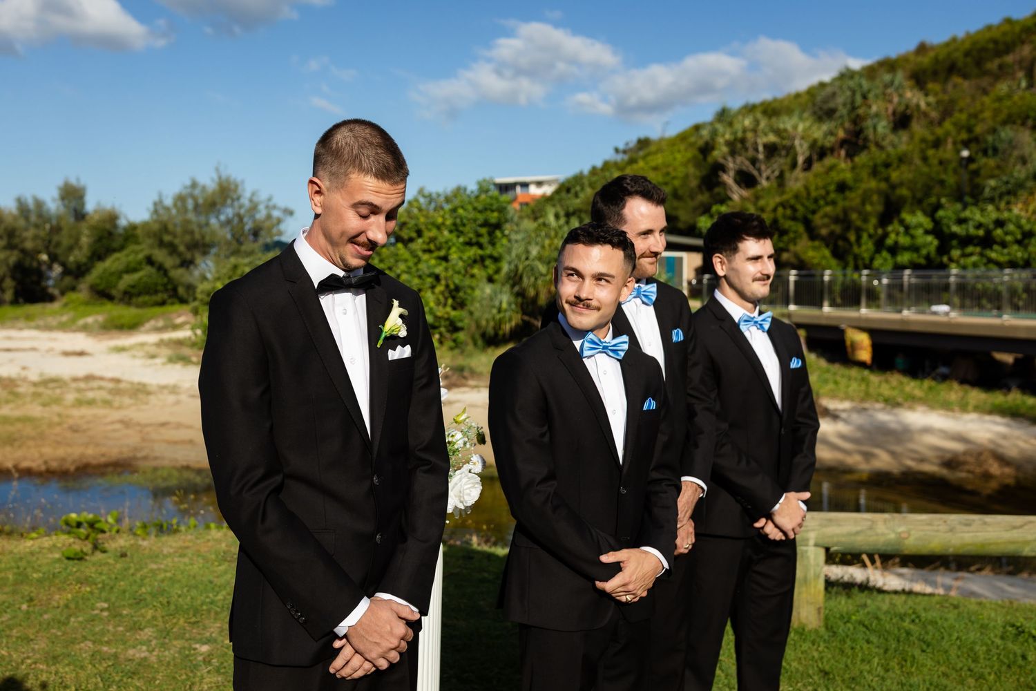 Groomsmen in black tuxedos pose together outdoors on a sunny day with green hills in the background.