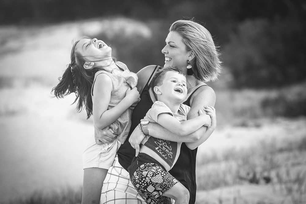 Black and white photo of a mother joyfully holding two children outdoors in a field.
