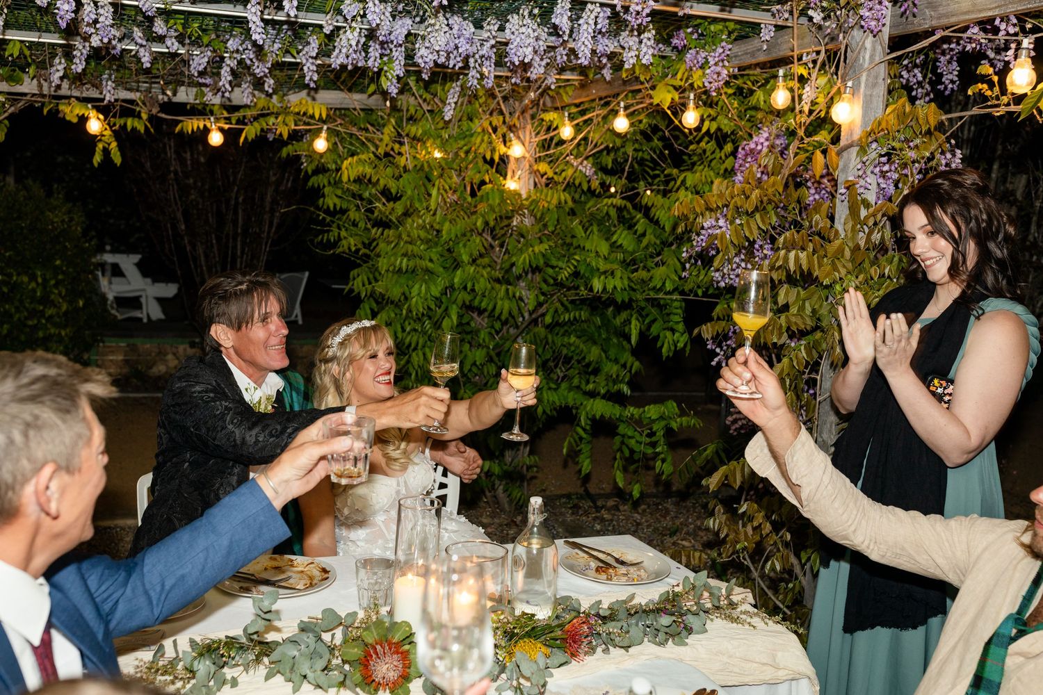 Guests celebrating with raised arms under string lights at an outdoor evening reception.