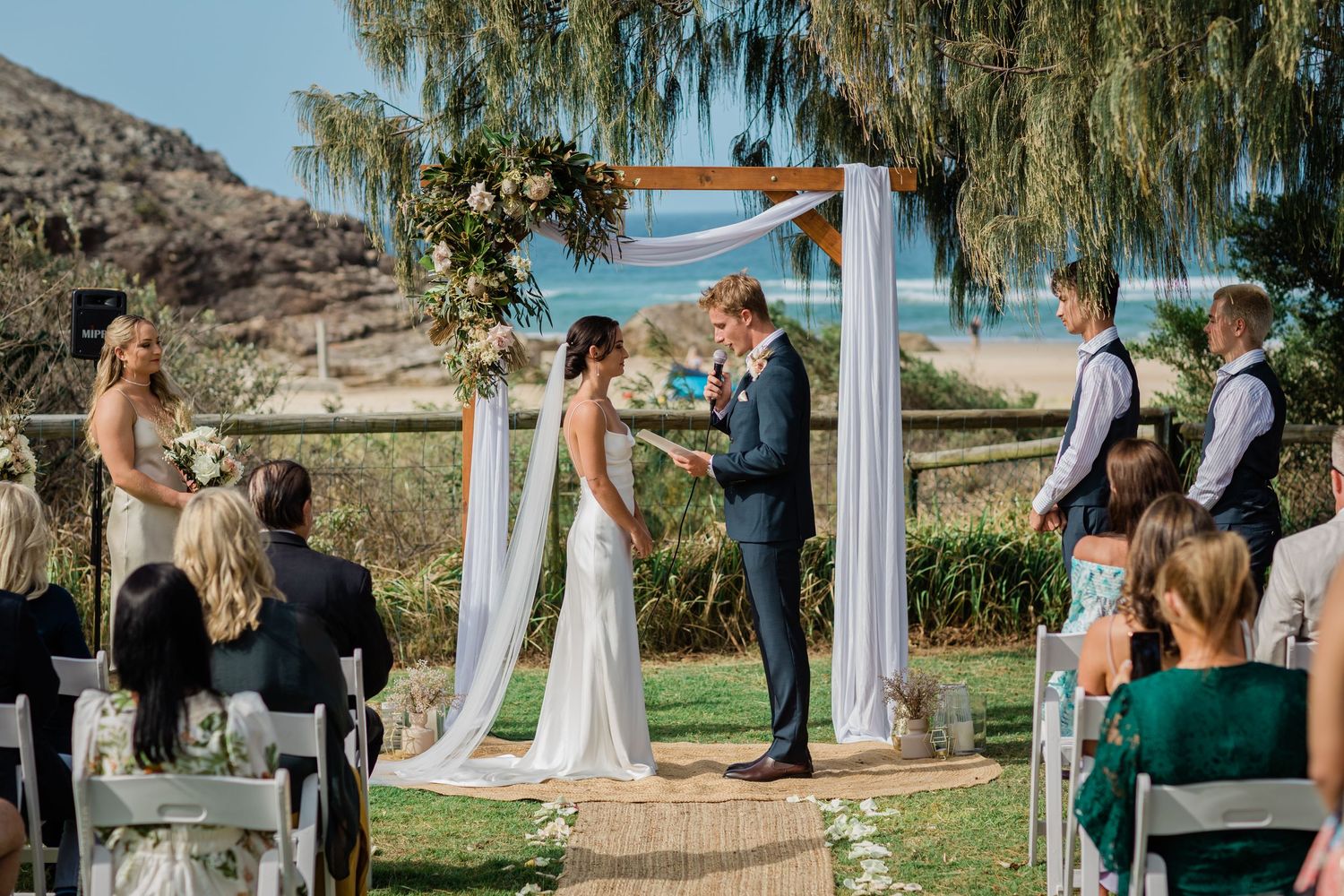 Wedding ceremony setup outdoors with white chairs and floral arch under willow trees.Miami Burleigh heads beach wedding