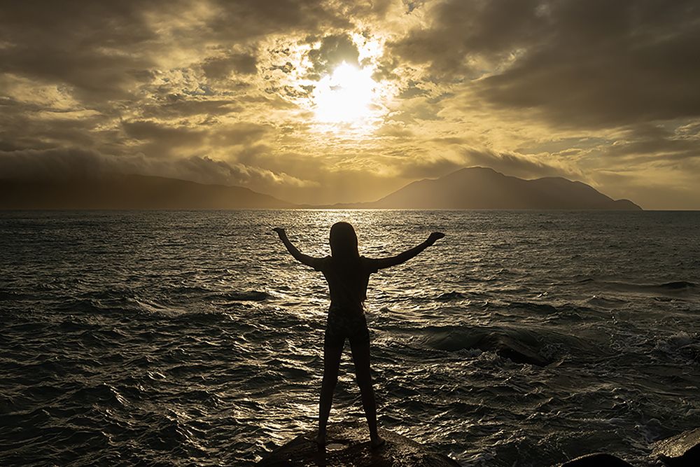 Silhouette of a person with raised arms against a dramatic sunset over the ocean.