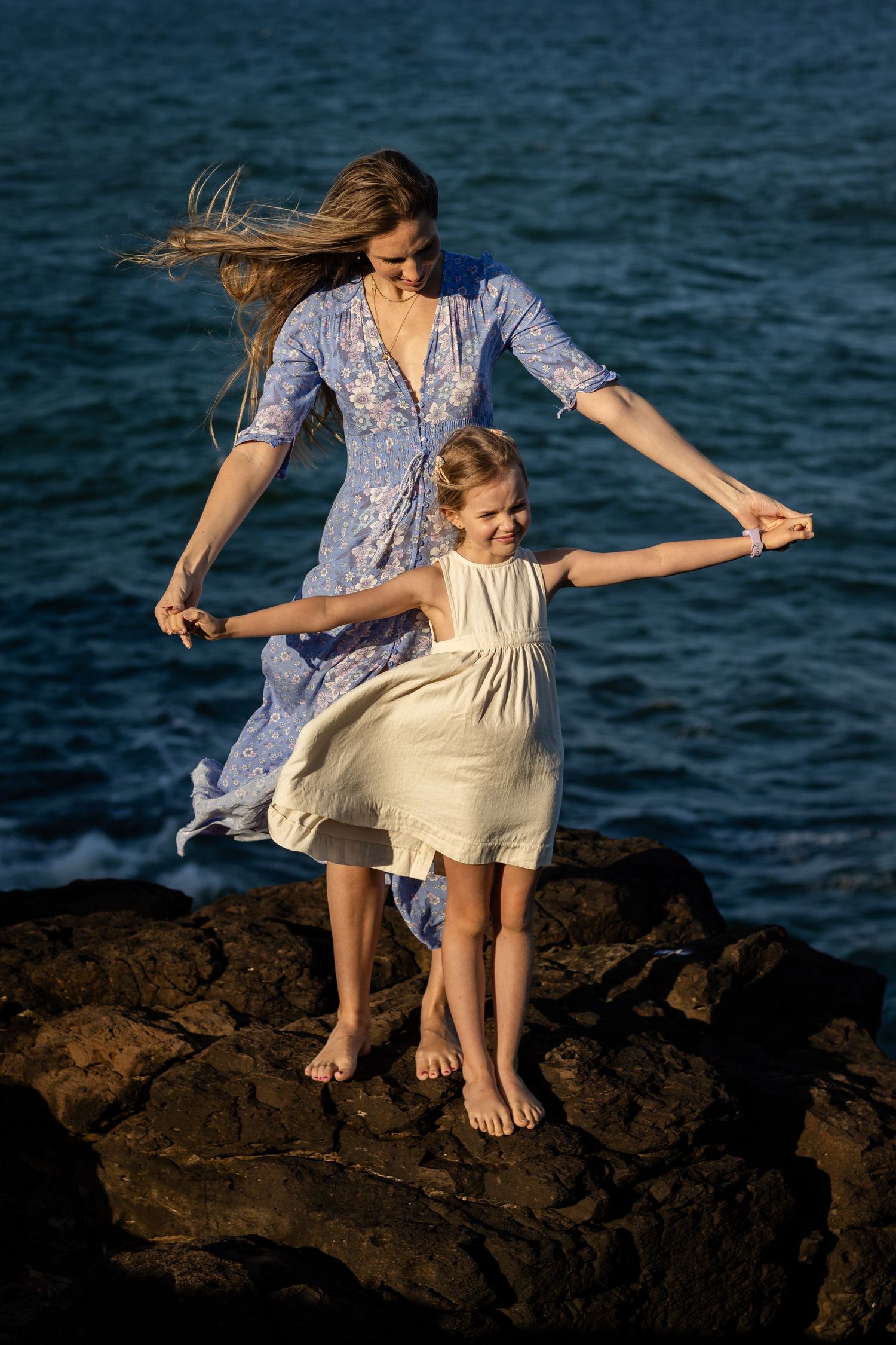 Two people in blue and cream dresses dance playfully on rocky coastal cliffs by the ocean.Gold Coast candid family photographer.
