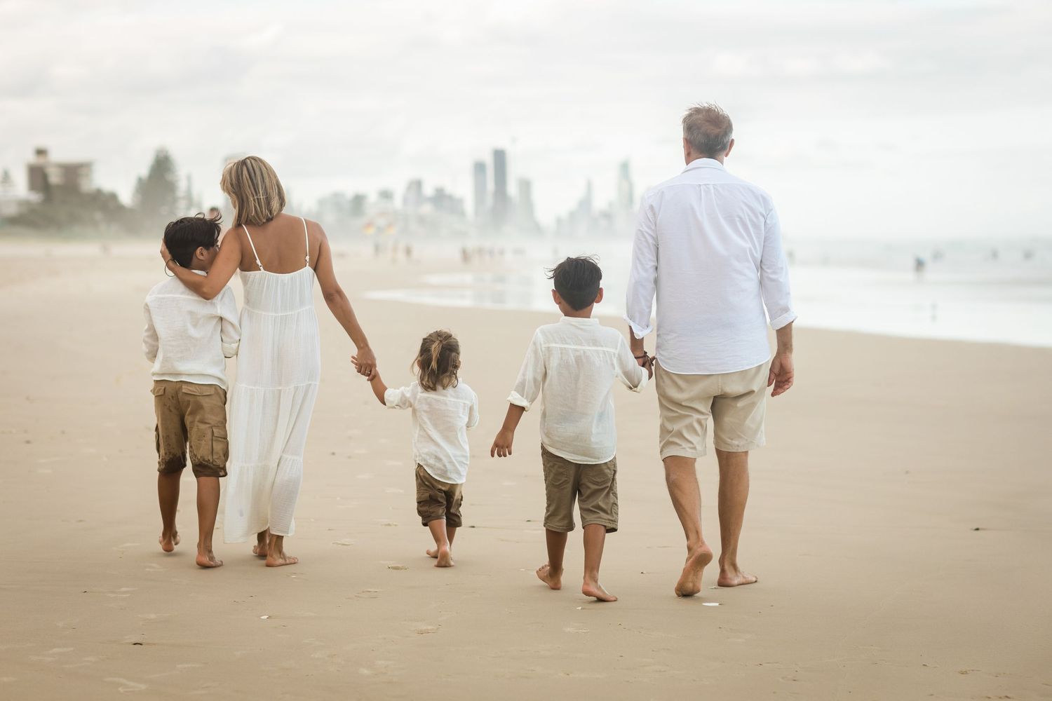Family walking together on sandy beach with distant cityscape visible.Gold Coast candid family photographer.