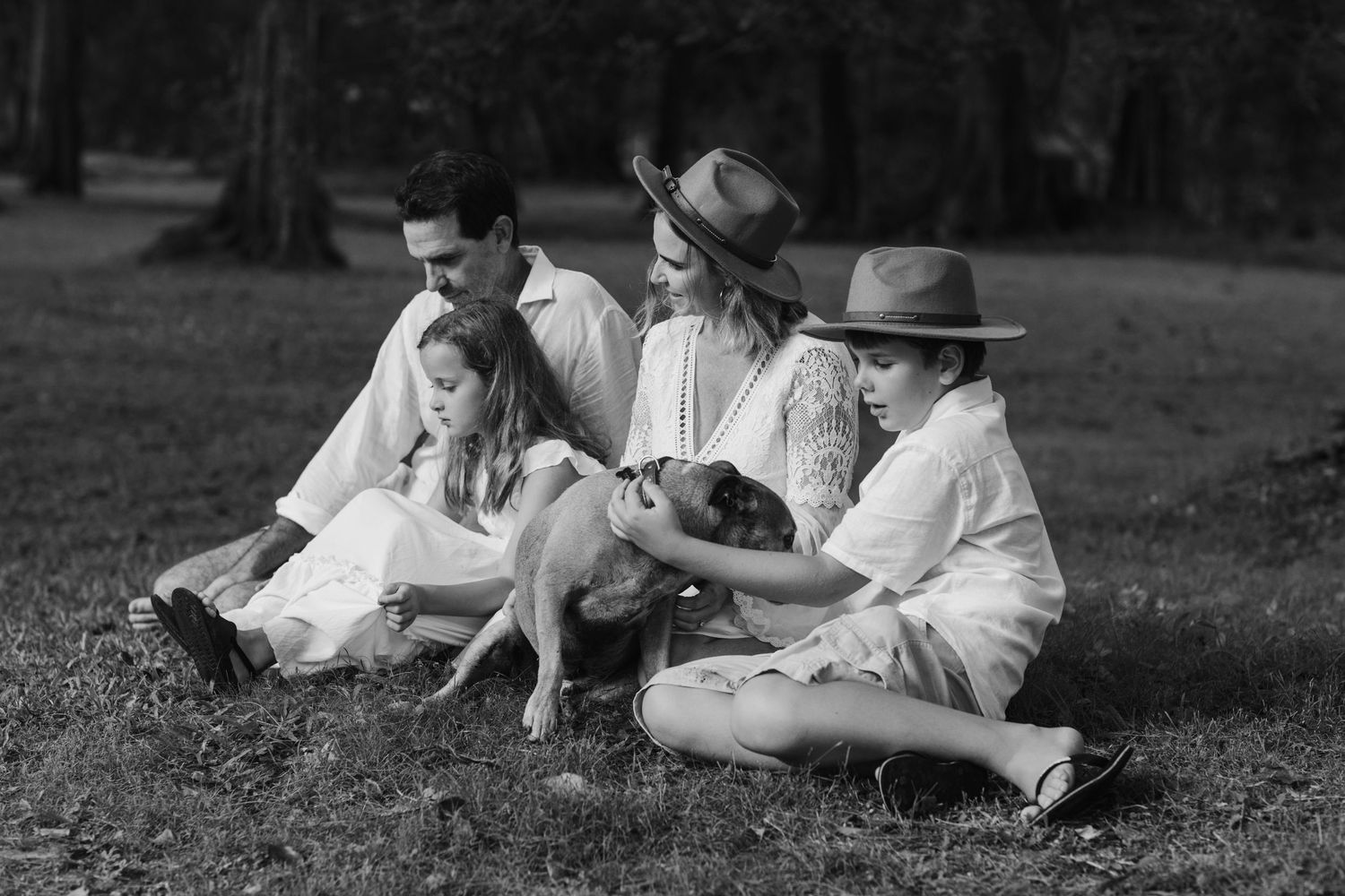 Family in white clothing sits together with their dog on grass in black and white.