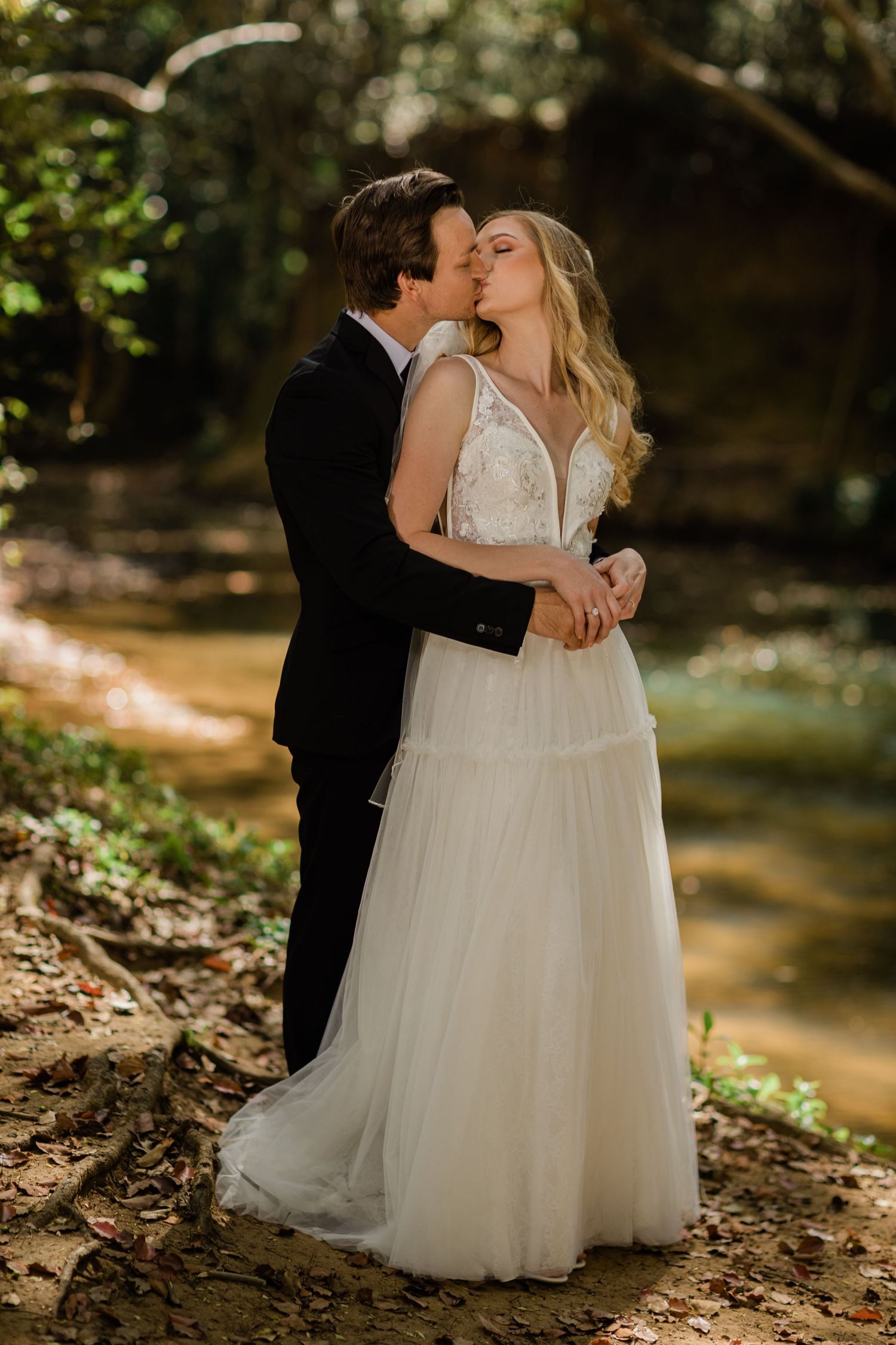 Newlyweds embrace on a forest path with sunlit creek in the background.Currumbin Valley wedding photography Gold Coast