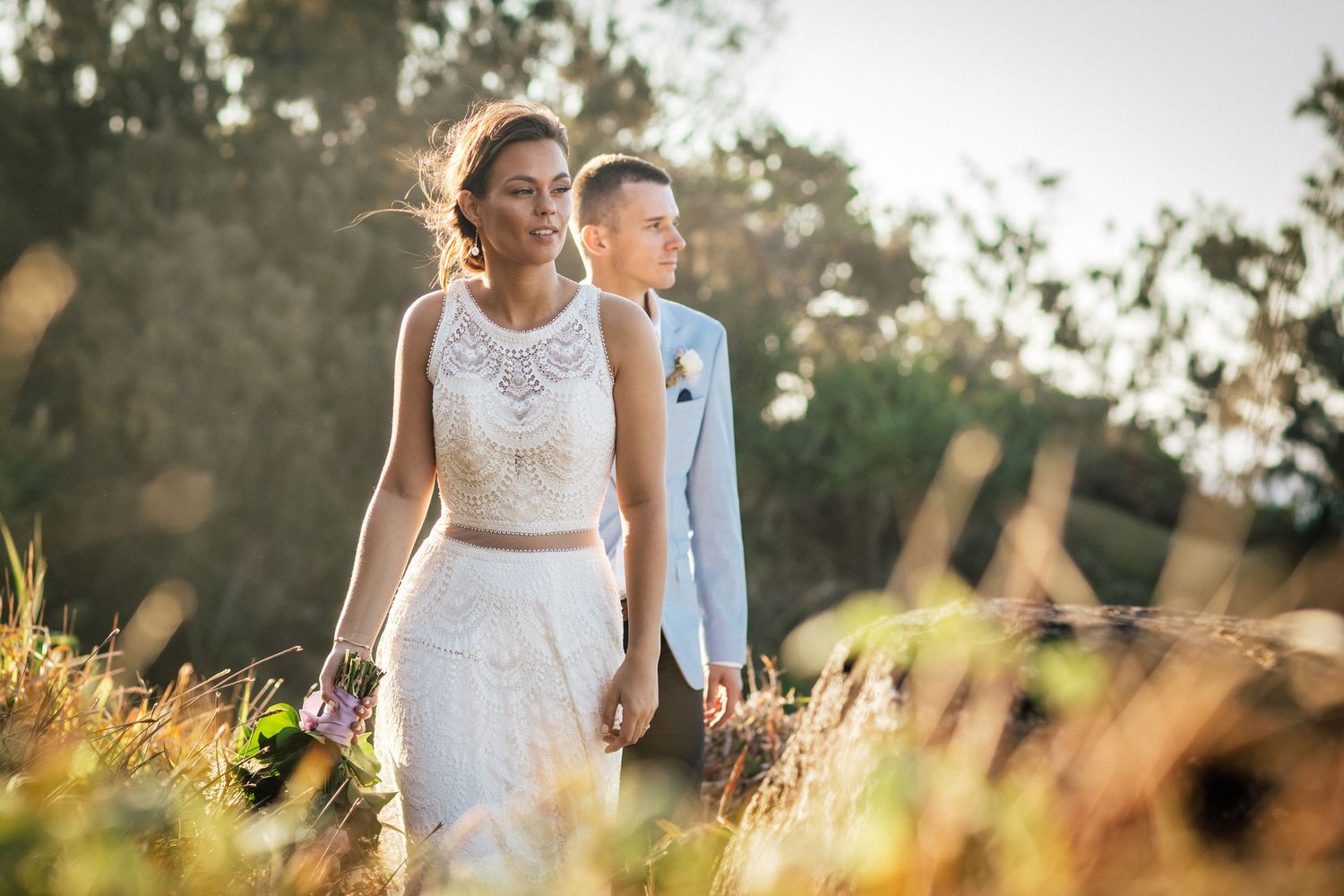 Couple walks through golden sunlit field during sunset wedding photo session.Gold Coast beach wedding photography. Froggies beach